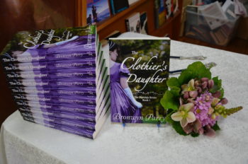 A table with a pile of copies of The Clothier's Daughter and a posy of flowers in mauve and white.