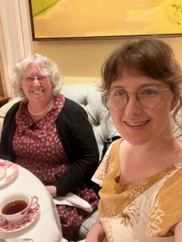 A young woman wearing a yellow and white dress with block-printed sunflowers seated at a table with an older woman wearing a maroon floral dress and a pearl nacklace.
