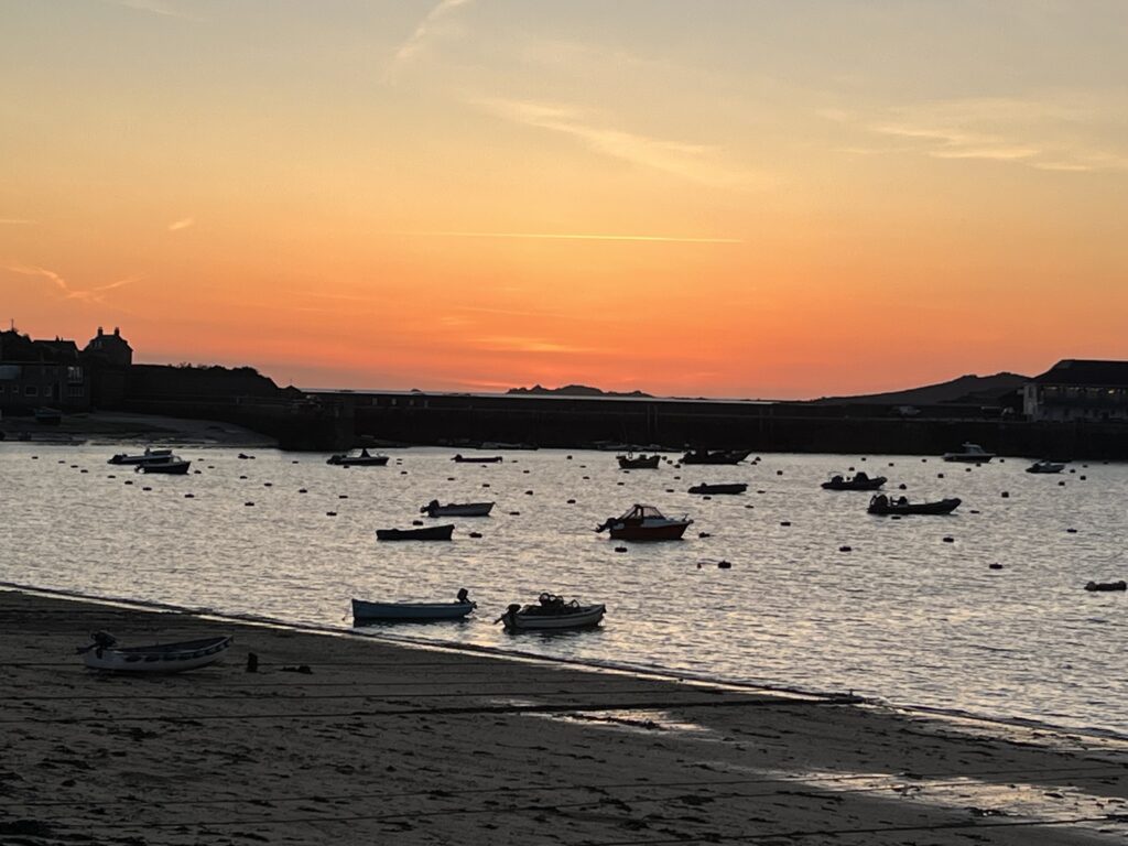 Red sky of sunset over the Hughtown bay, with dark shapes of boats visible on the calm, grey sea.